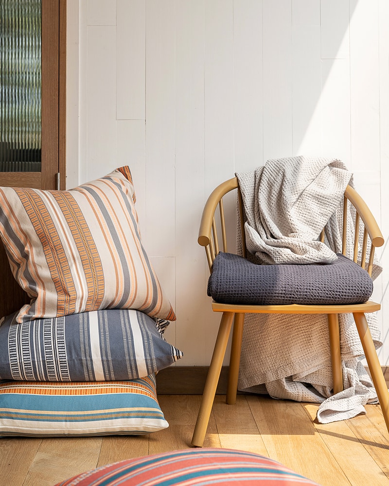 A stack of Mungo Sahara Stripe Cushion Covers next to the Mungo Cobble Weave Bed Cover