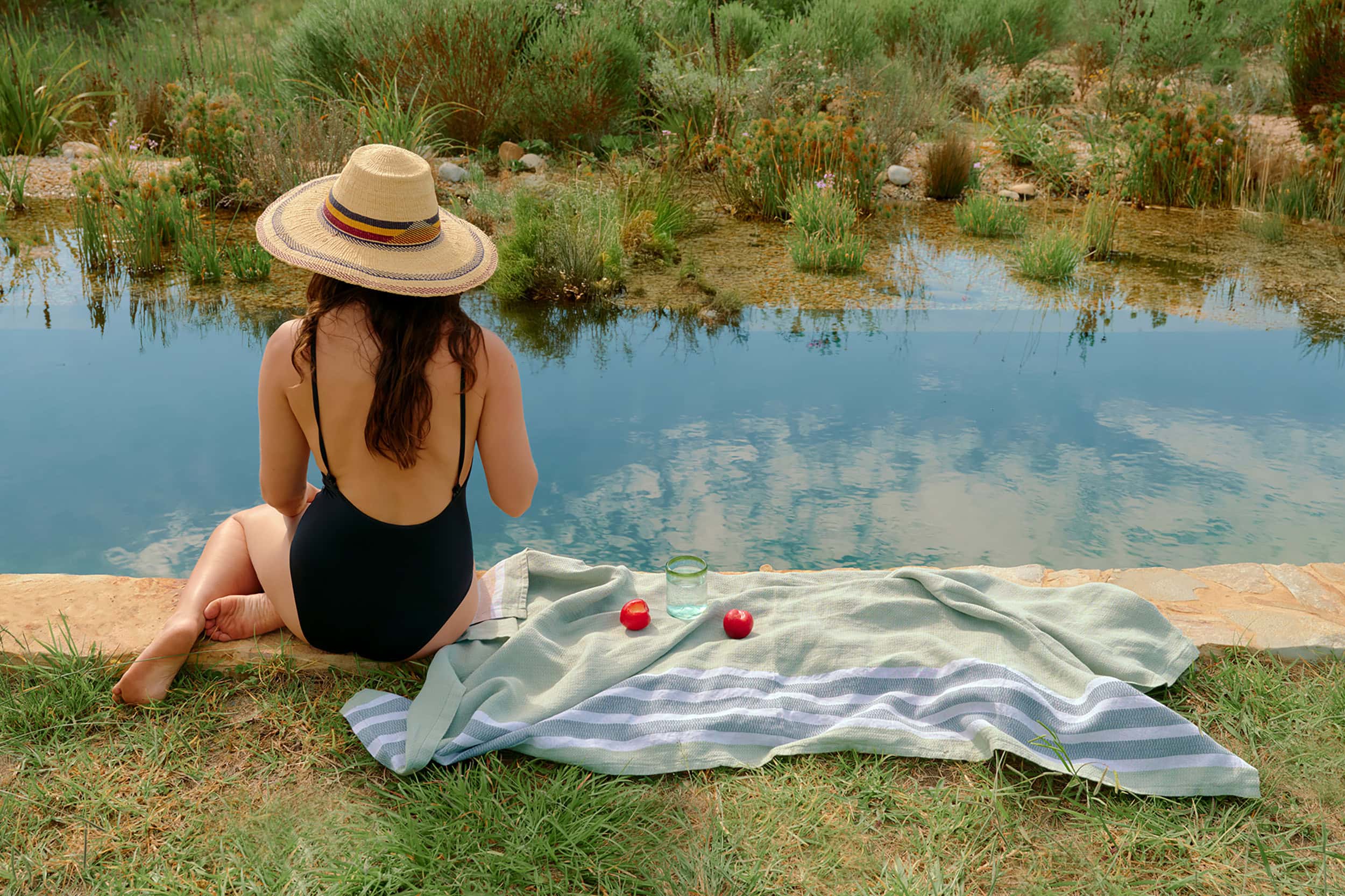 Model, wearing a hat, sitting by a natural pool with a Mungo flax towel.